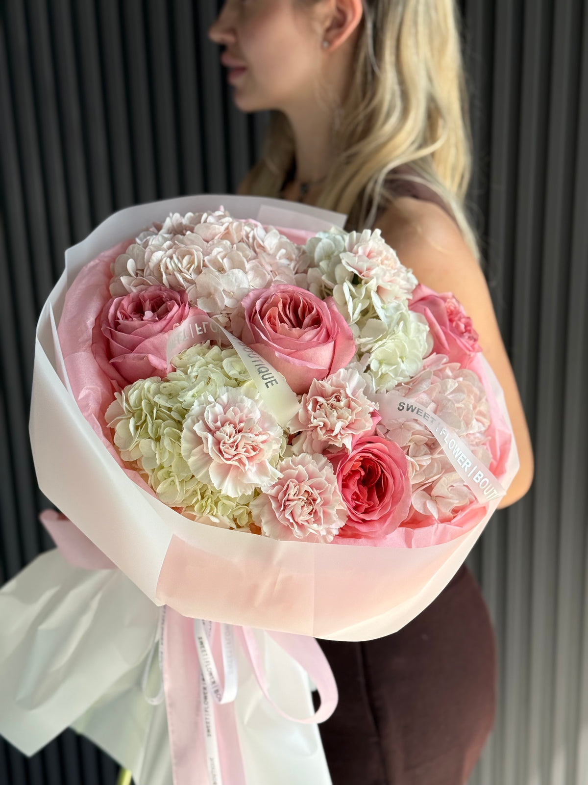 A bouquet of pink and white flowers wrapped in pale pink tissue paper and white paper, held by a woman in a dark brown dress against a dark gray slatted wall.