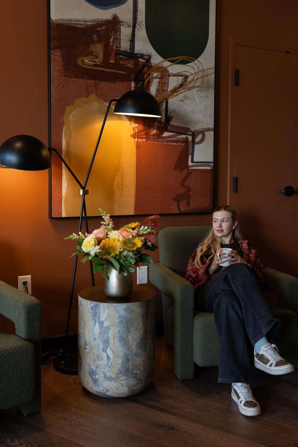 A woman sits in a green armchair in a warm, modern living room, holding a mug. She is wearing a red patterned shirt, dark wide-leg pants, and white sneakers.