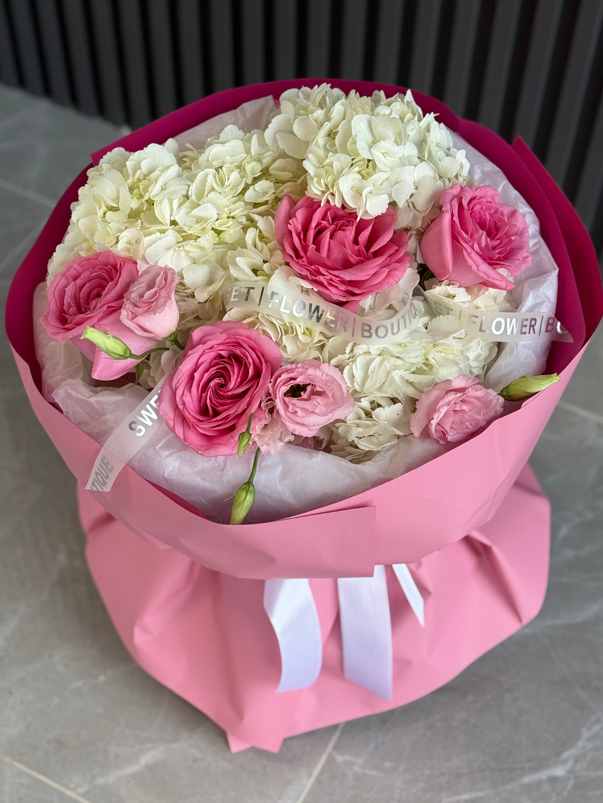 Bouquet of white and pink flowers wrapped in pink paper with a visible brand label on a gray surface.