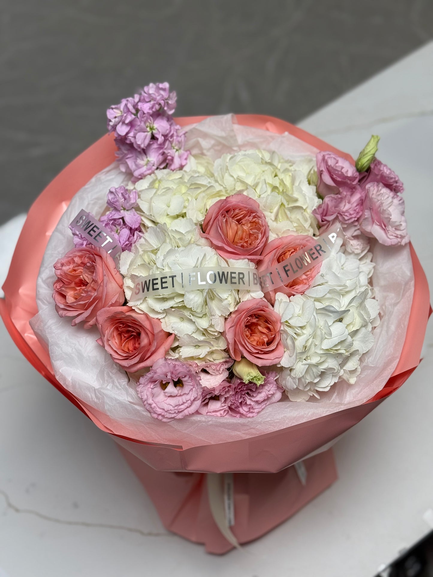 Bouquet of flowers wrapped in pink paper with visible brand name on a white surface.