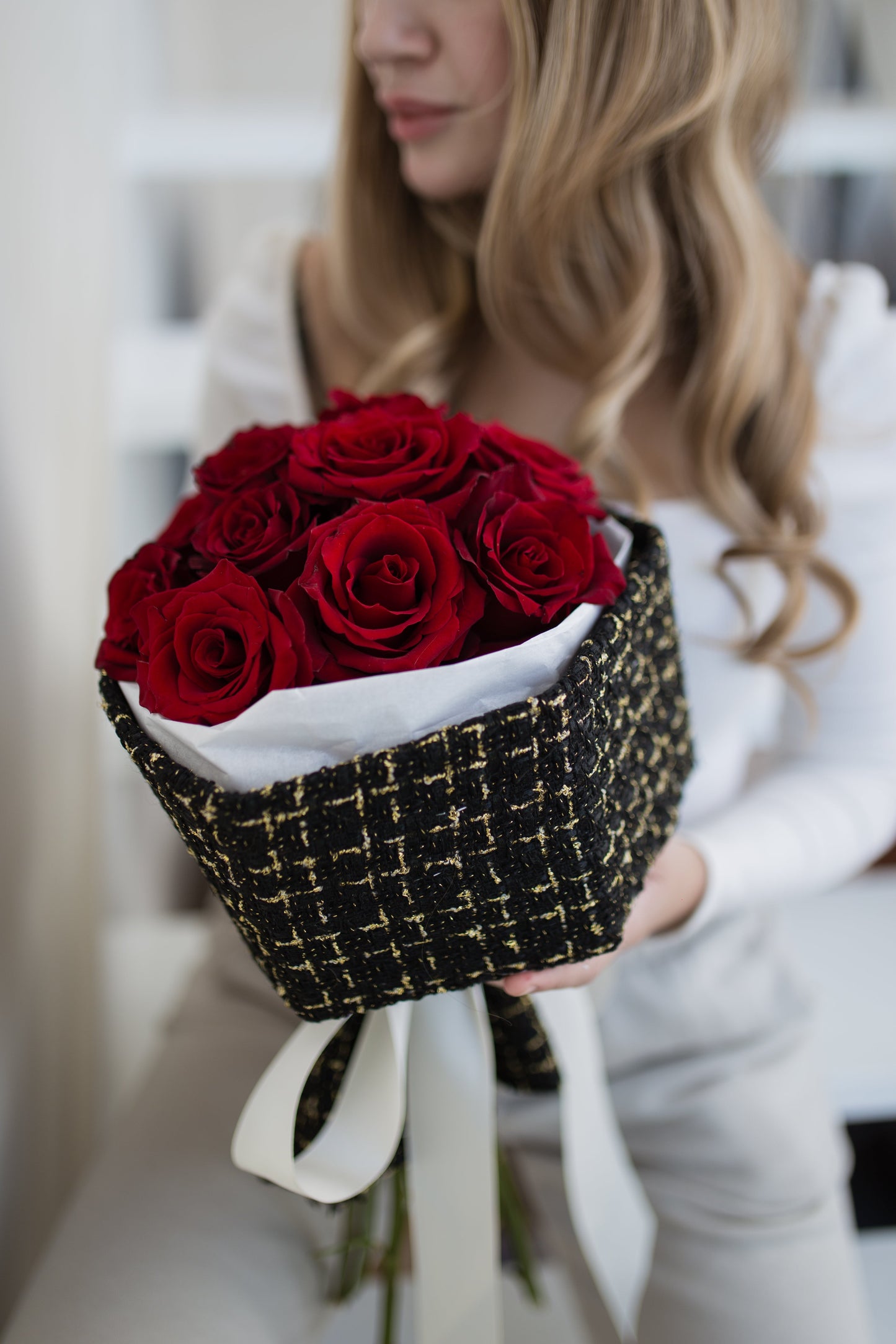 A person holding a bouquet of red roses wrapped in black and gold tweed fabric.
