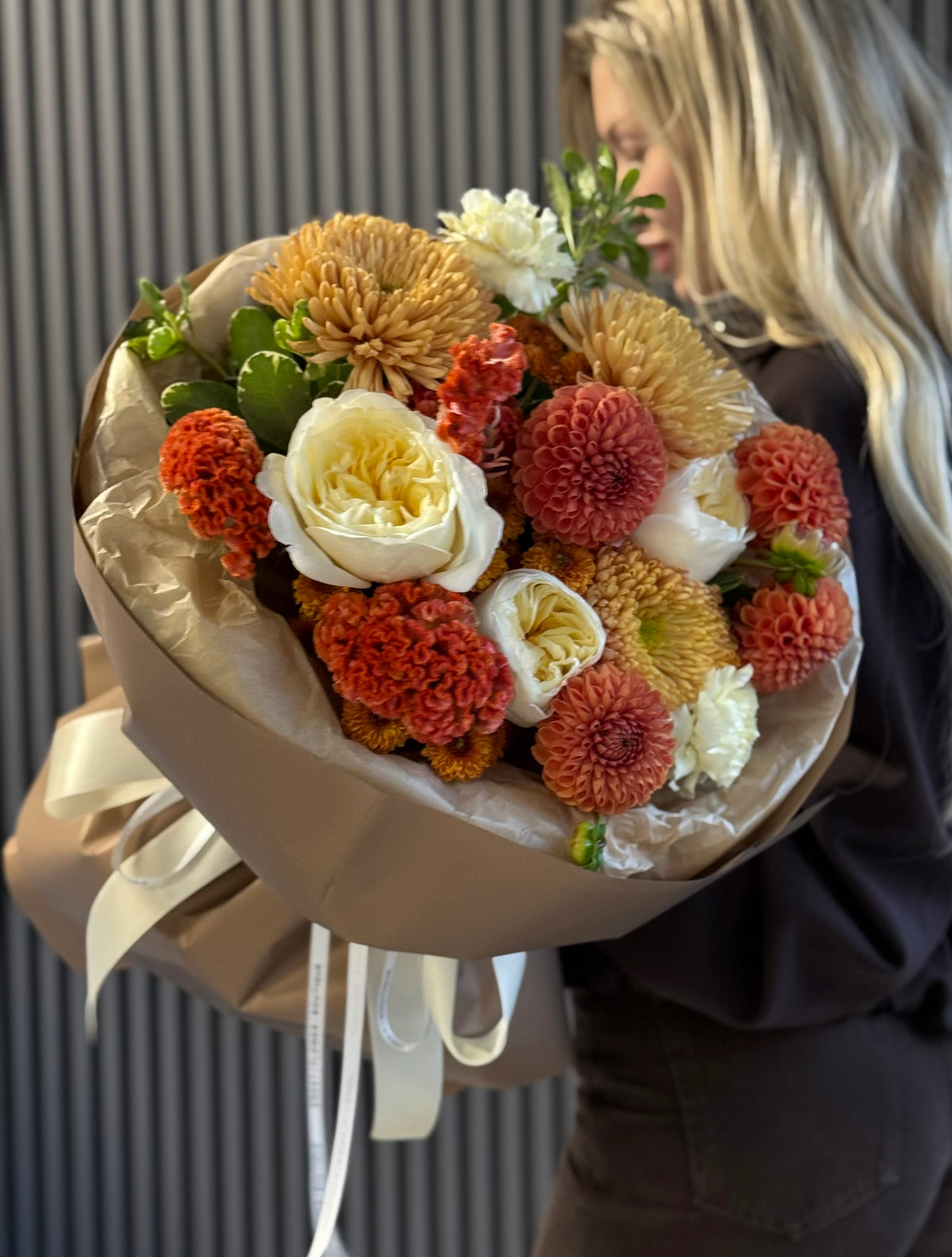 A large, beautifully arranged floral bouquet with orange, red, cream, and yellow flowers wrapped in brown paper with a white ribbon, held by a woman with blonde hair against a dark gray paneled wall.