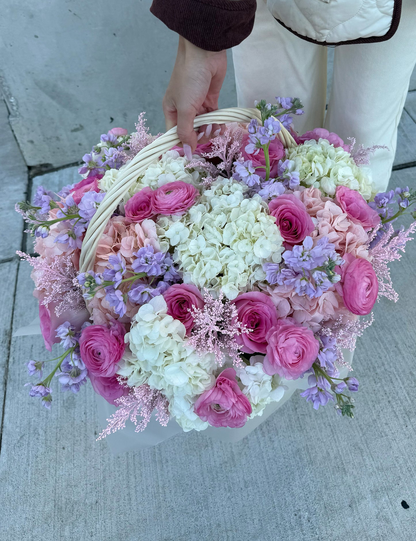 Basket of pink, white, and purple flowers held by a person on a concrete surface.