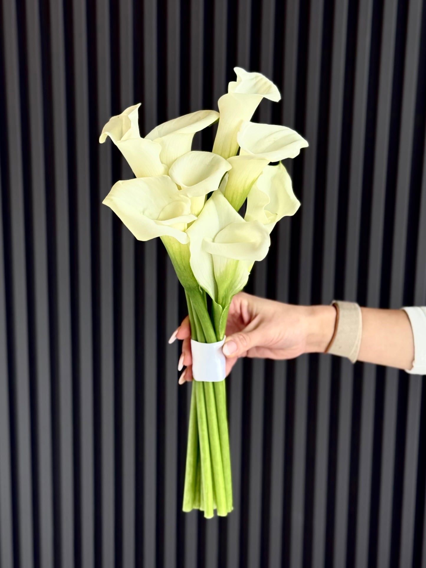 A hand holding a bouquet of white calla lilies with green stems against a dark gray vertically slatted background.