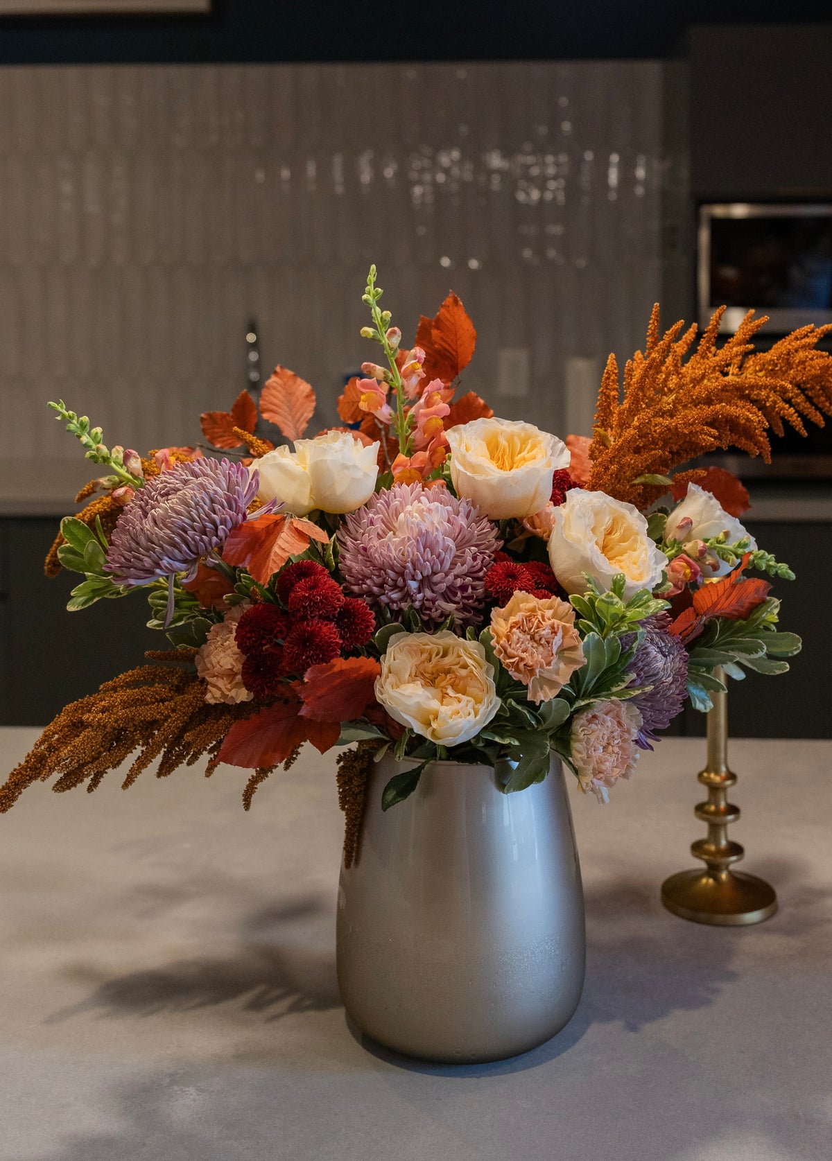 An autumn floral arrangement in a matte silver vase, featuring orange and red foliage, purple and cream roses, and green leaves, set on a countertop with a blurred kitchen background.