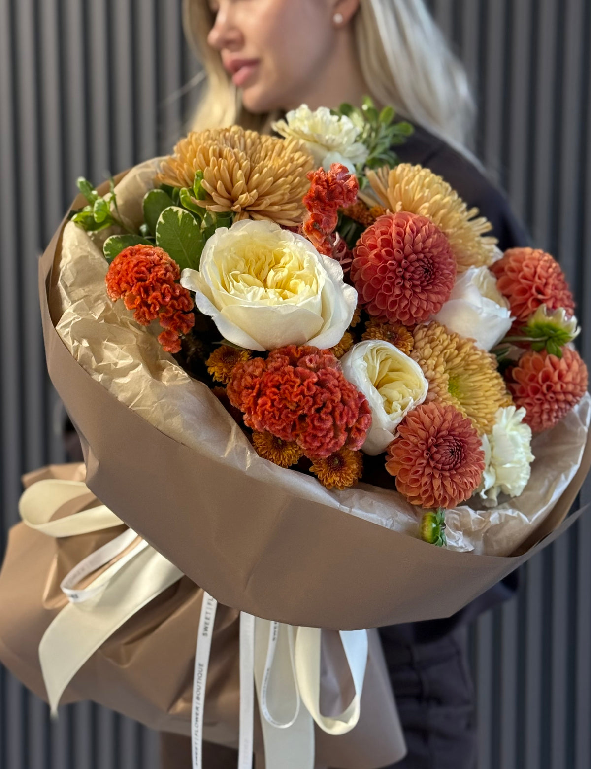 A woman holds a large, beautifully arranged autumn flower bouquet wrapped in brown paper with a cream ribbon. The bouquet features a mix of orange, red, yellow, and white flowers, including chrysanthemums, dahlias, and a white rose, set against a dark gray paneled background.