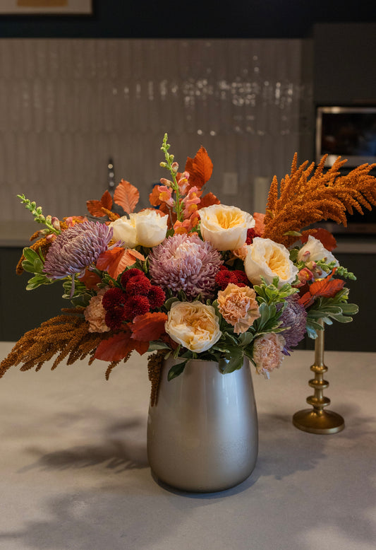 An autumnal floral arrangement in a matte silver vase, featuring orange, red, purple, and cream flowers with green foliage, set on a countertop in a modern kitchen.