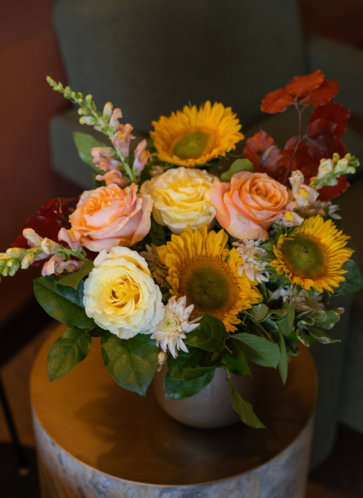 A vibrant autumn floral arrangement featuring yellow sunflowers, peach and cream roses, and red poppies with green foliage, presented in a neutral-toned ceramic pot on a rustic wooden side table.