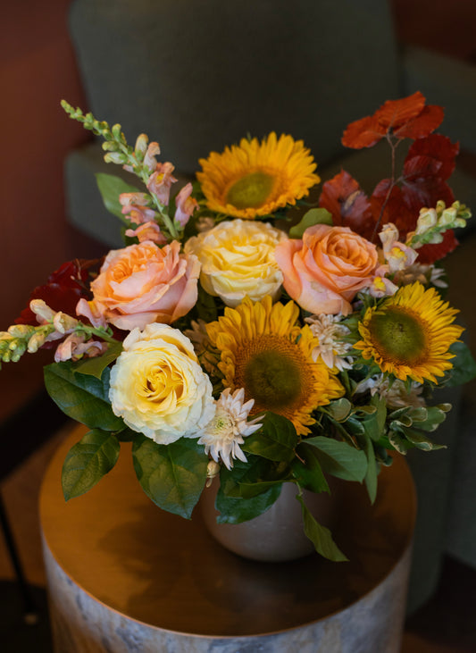 A vibrant autumn floral arrangement featuring yellow sunflowers, peach and cream roses, and red poppies with green foliage, presented in a neutral-toned ceramic pot on a rustic wooden side table.