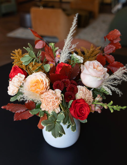 A floral arrangement in a white ceramic vase, featuring red and peach roses, carnations, variegated green foliage, and dried ornamental grasses on a dark tabletop.