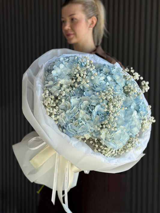 A woman holding a large bouquet of dried light blue hydrangeas and white baby's breath flowers, wrapped in white tissue paper with a cream ribbon, against a dark paneled background.