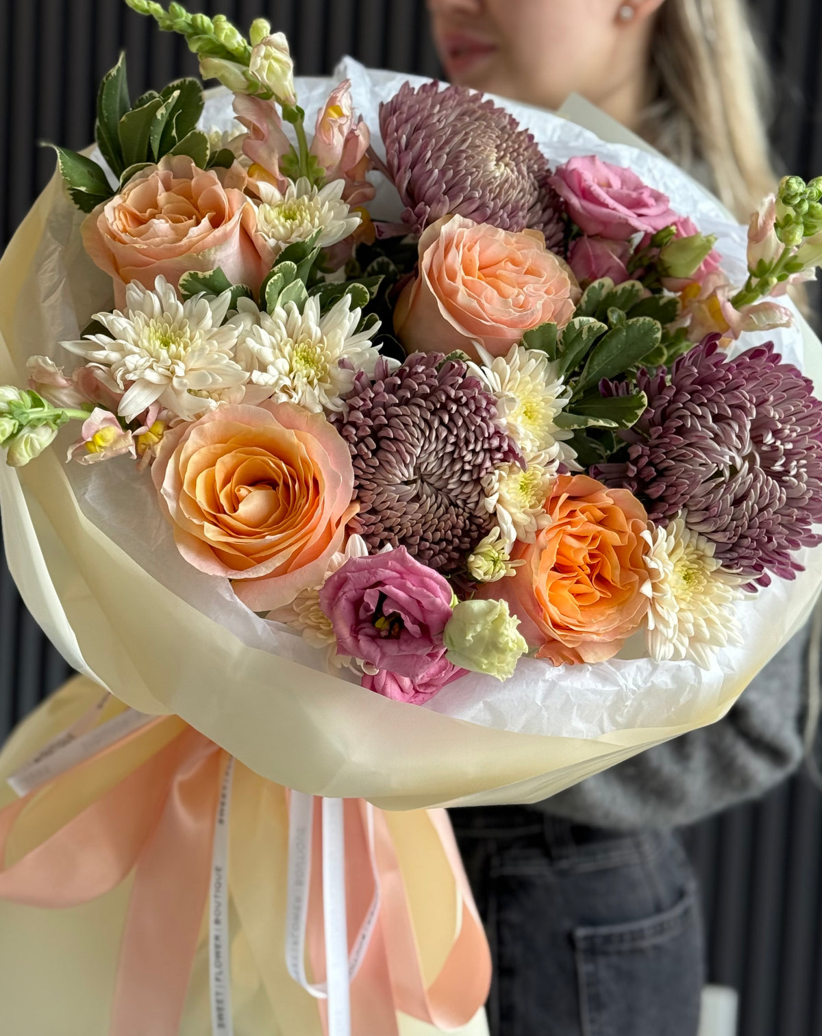A large, beautifully arranged floral bouquet held by a person. The bouquet features a mix of peach roses, deep purple chrysanthemums, white daisies, and pink lisianthus wrapped in cream paper with a peach ribbon.