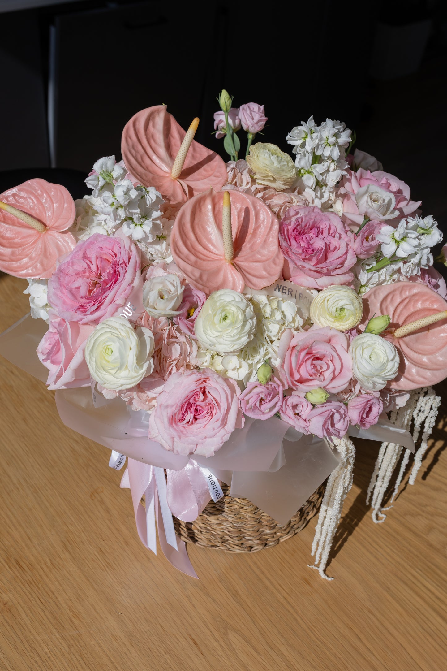 A large, vibrant floral arrangement in a woven basket, featuring pink and white flowers including anthuriums, roses, ranunculus, and hydrangeas, wrapped in pale pink paper with a ribbon on a wooden table.