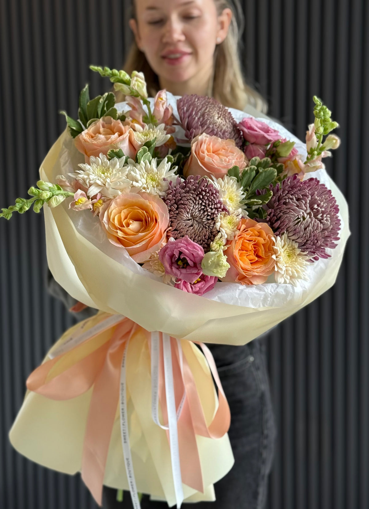 A woman holding a large, beautifully arranged bouquet of fresh flowers. The bouquet features peach roses, orange roses, purple chrysanthemums, and white chrysanthemums, wrapped in cream paper with peach and white ribbons.