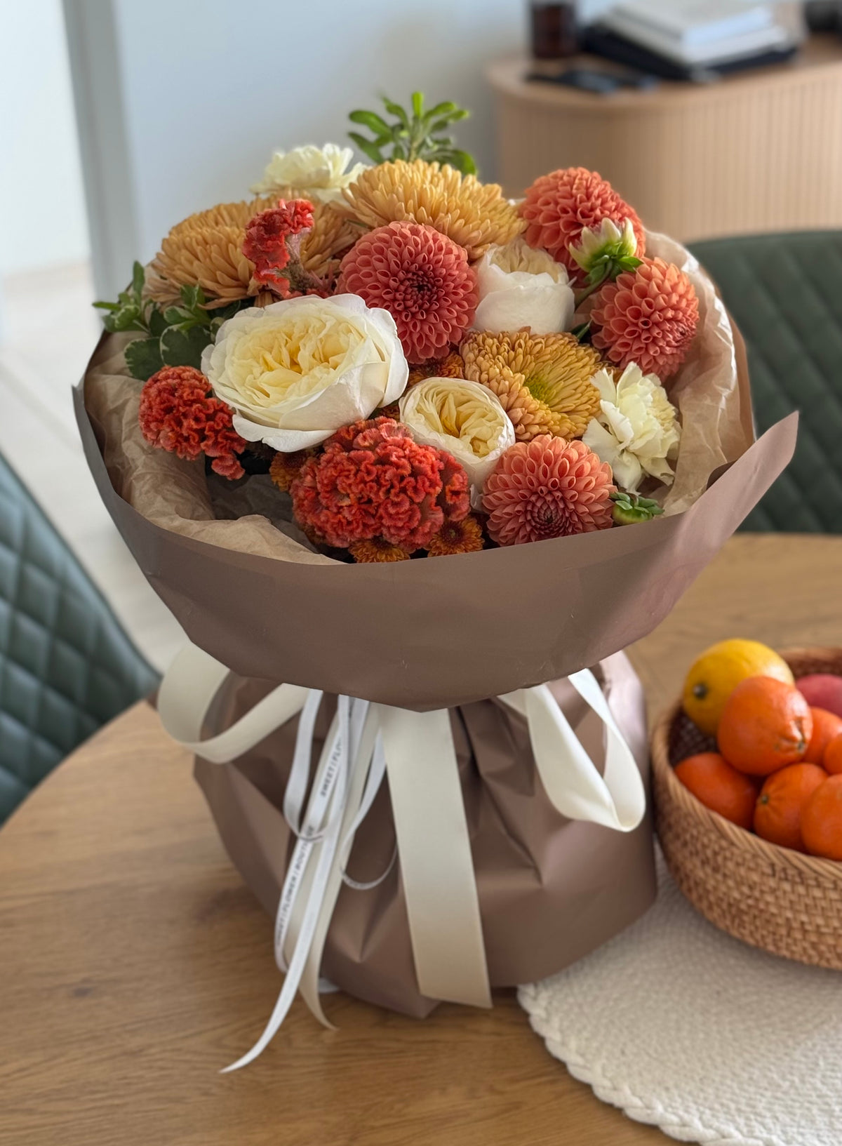 A wrapped bouquet of orange, peach, cream, and yellow flowers in brown paper wrapping with a white ribbon, placed on a wooden table next to a basket of oranges.