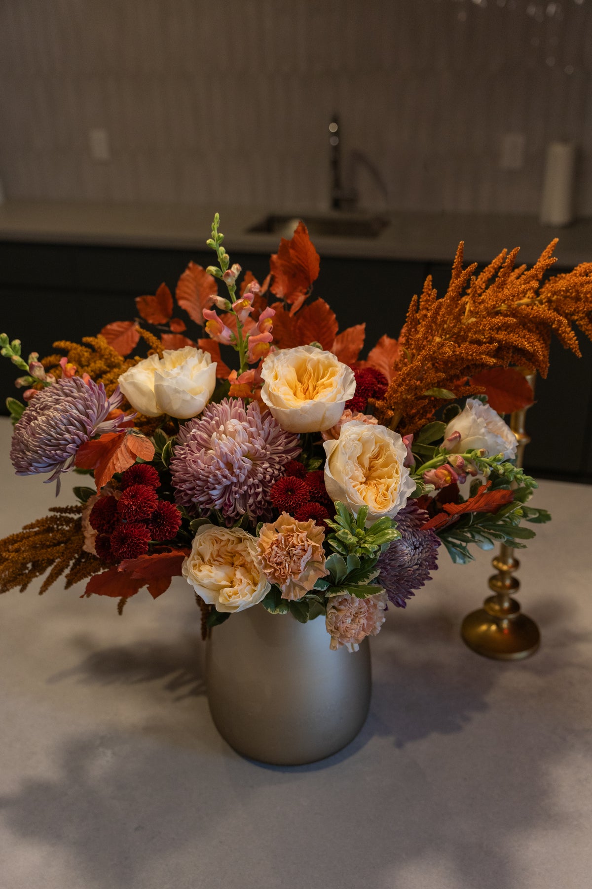 An autumnal floral arrangement in a matte silver vase, featuring cream roses, burgundy chrysanthemums, orange foliage, and purple flowers on a kitchen counter.