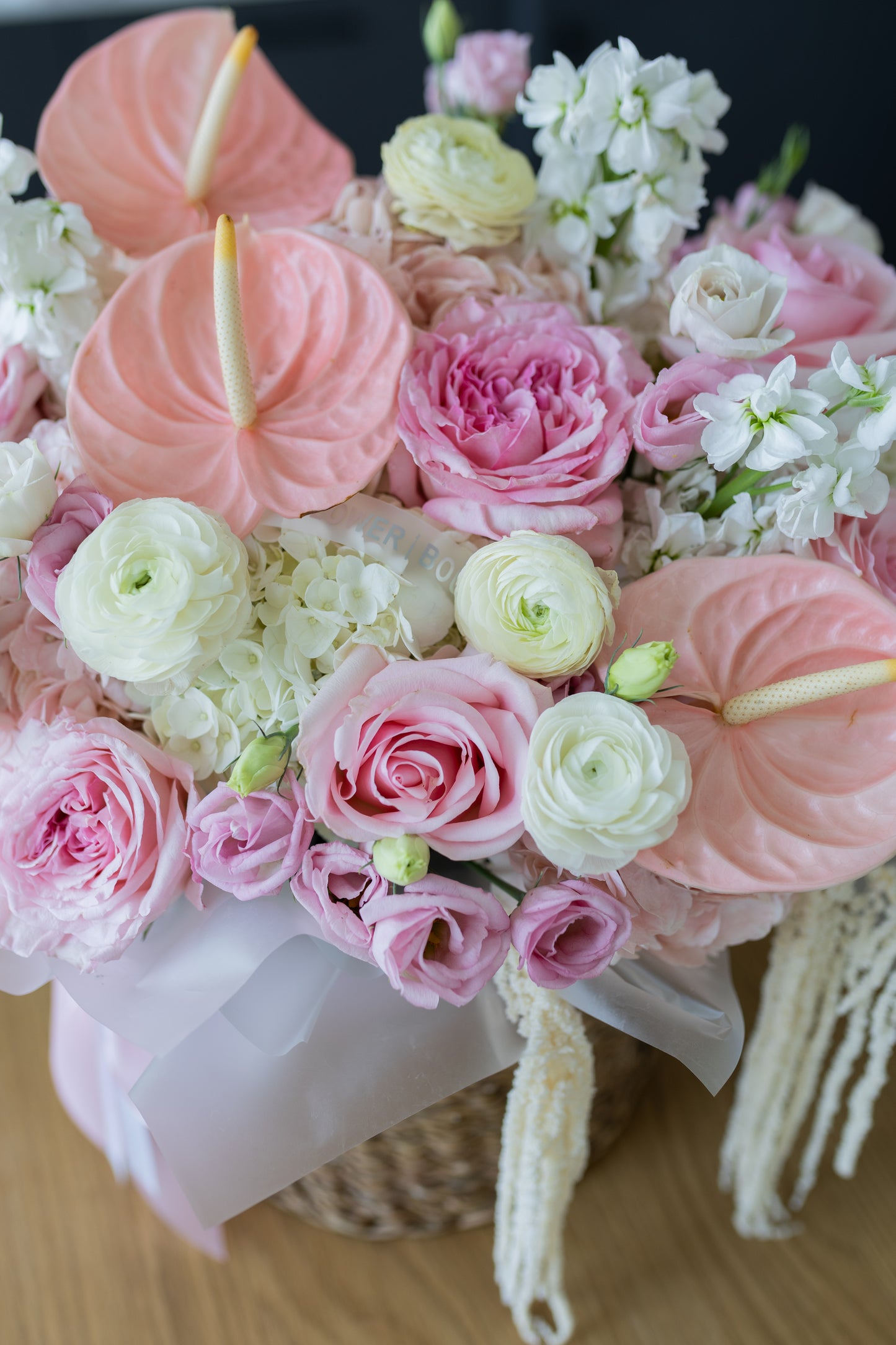 A close-up of a floral arrangement featuring pink and white flowers, including anthuriums, roses, ranunculus, and hydrangeas, tied with a pale pink ribbon and hanging tassels, set against a dark background.