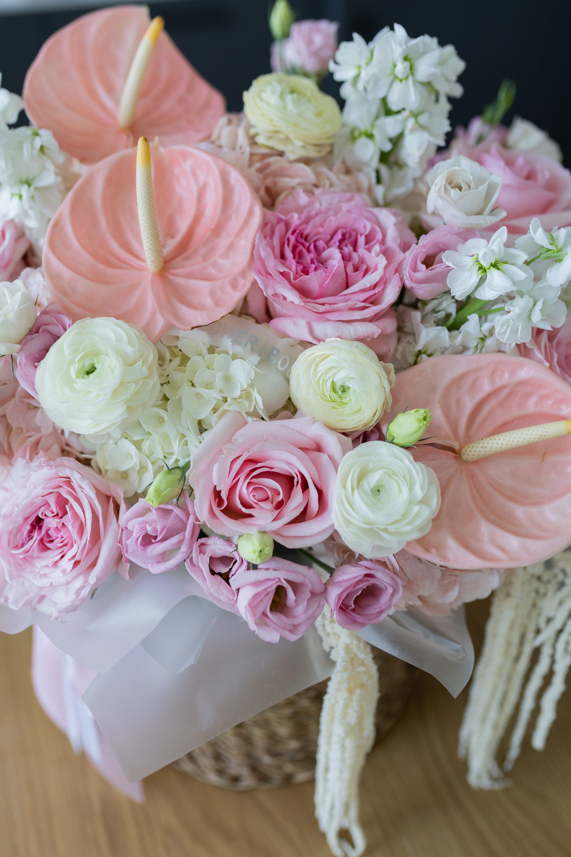 A close-up of a floral arrangement featuring pink and white flowers, including anthuriums, roses, ranunculus, and hydrangeas, tied with a pale pink ribbon and hanging tassels, set against a dark background.