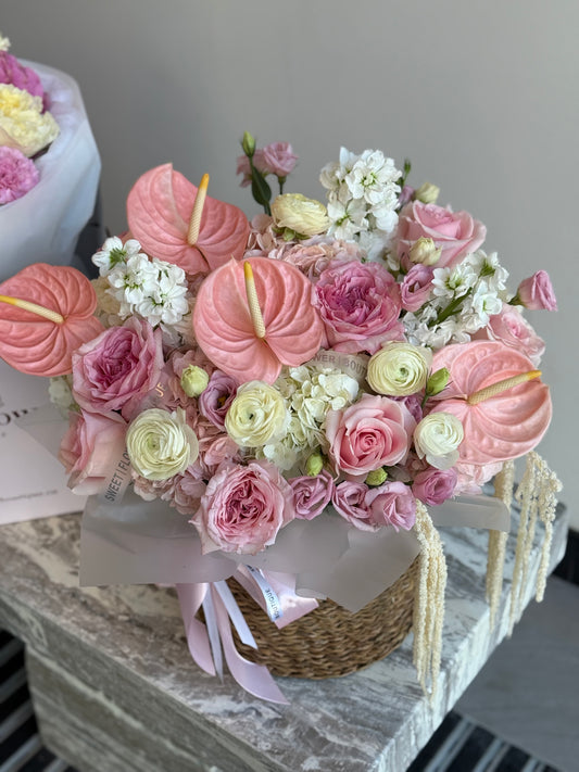 A large, vibrant floral arrangement featuring pink and white flowers in a woven basket with a pink ribbon, displayed on a marble surface.