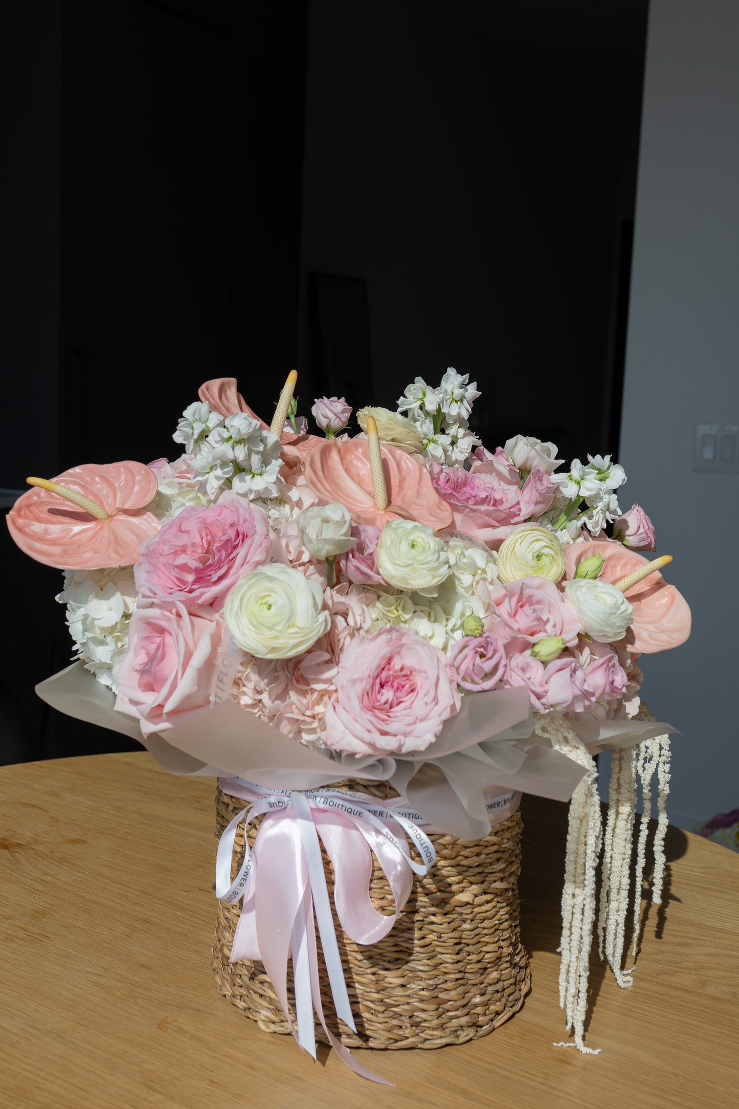 A large floral arrangement in a woven basket with pink and white flowers, wrapped in white tissue paper and tied with a pale pink ribbon, sitting on a wooden table.