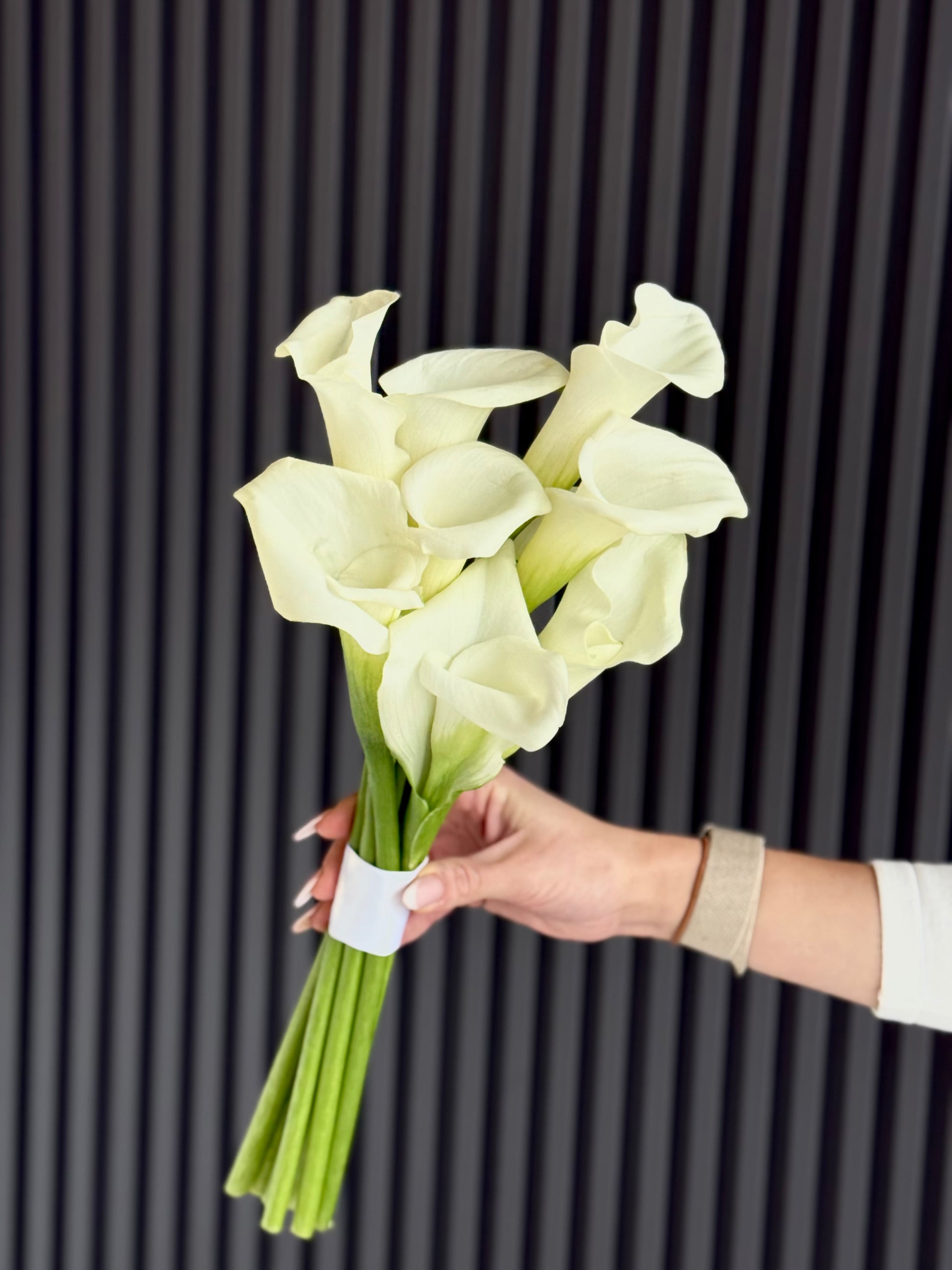 A bouquet of white calla lilies with green stems held by a person's hand against a dark gray vertically slatted background.