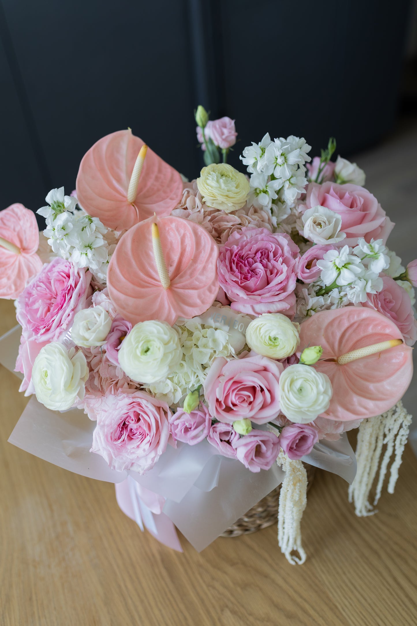 A vibrant floral arrangement featuring pink and white flowers, including anthuriums, roses, ranunculus, and white flowers, wrapped in pale pink paper and tied with a ribbon, sitting on a light wooden surface.
