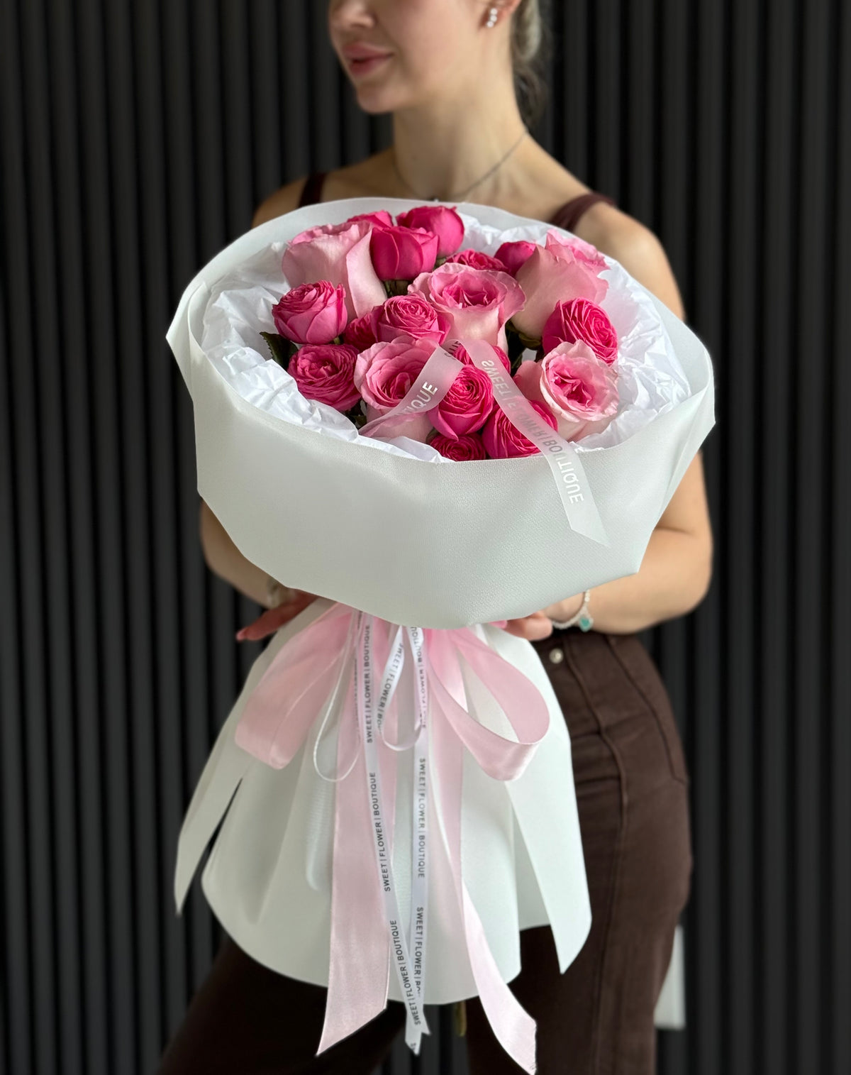 A bouquet of pink roses wrapped in white paper with a light pink ribbon, held by a woman against a dark vertical slat background.