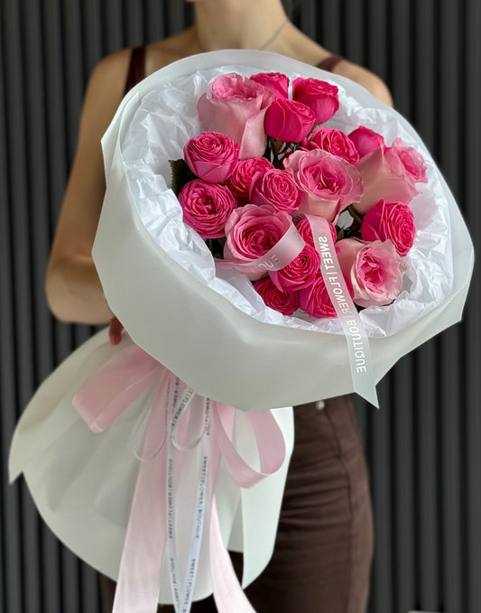 A person holding a large bouquet of pink roses wrapped in white paper with a light pink ribbon, against a dark gray paneled background.