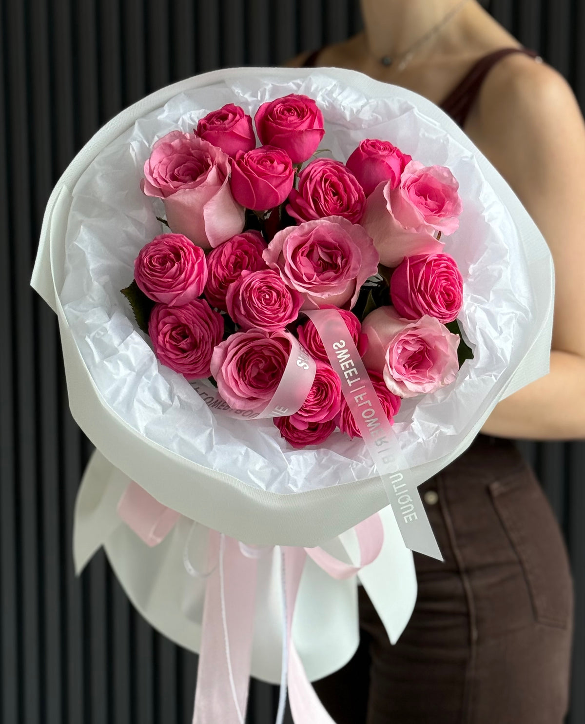 A bouquet of pink roses wrapped in white tissue paper and a white paper sleeve with pink ribbon ties, held by a person wearing a brown top and brown pants against a dark gray paneled wall.