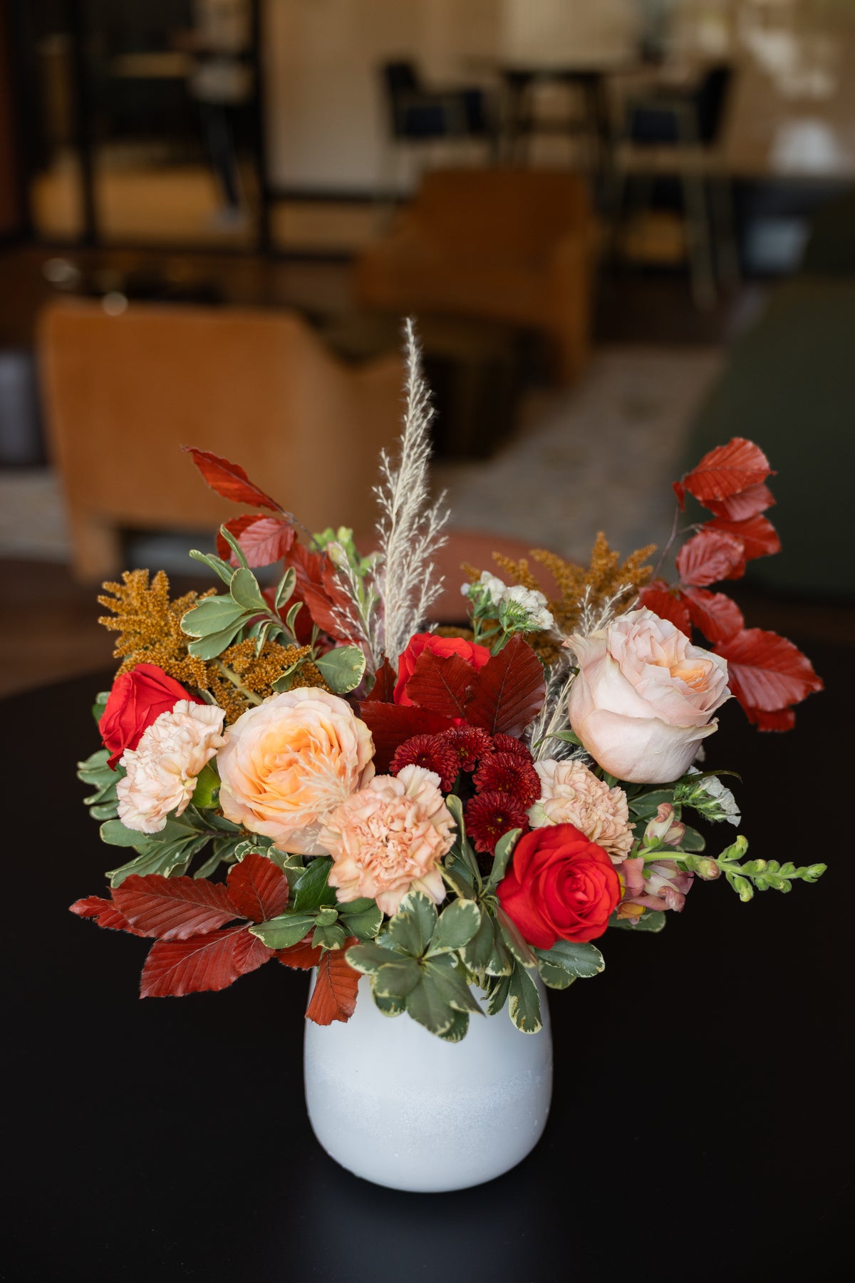 A vibrant autumn floral arrangement in a white ceramic vase, featuring red and peach roses, burgundy carnations, variegated green foliage, and warm orange-brown leaves, set against a blurred indoor background.