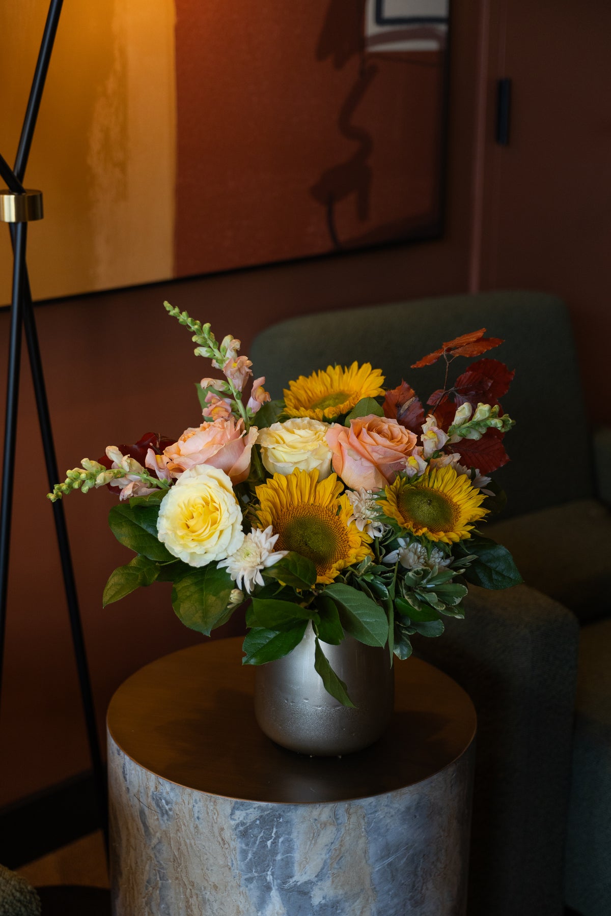 A vibrant floral arrangement featuring yellow sunflowers, peach roses, and white daisies in a metallic silver vase, displayed on a round wooden side table in a cozy, warmly lit living room setting.