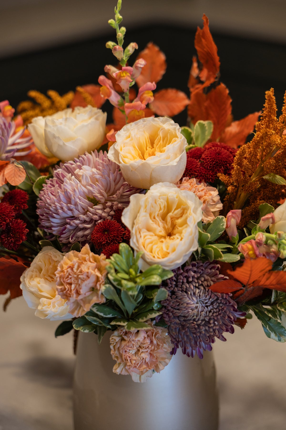 A close-up of a floral arrangement in a white vase, featuring cream roses, purple chrysanthemums, red mums, orange autumn leaves, and green foliage.