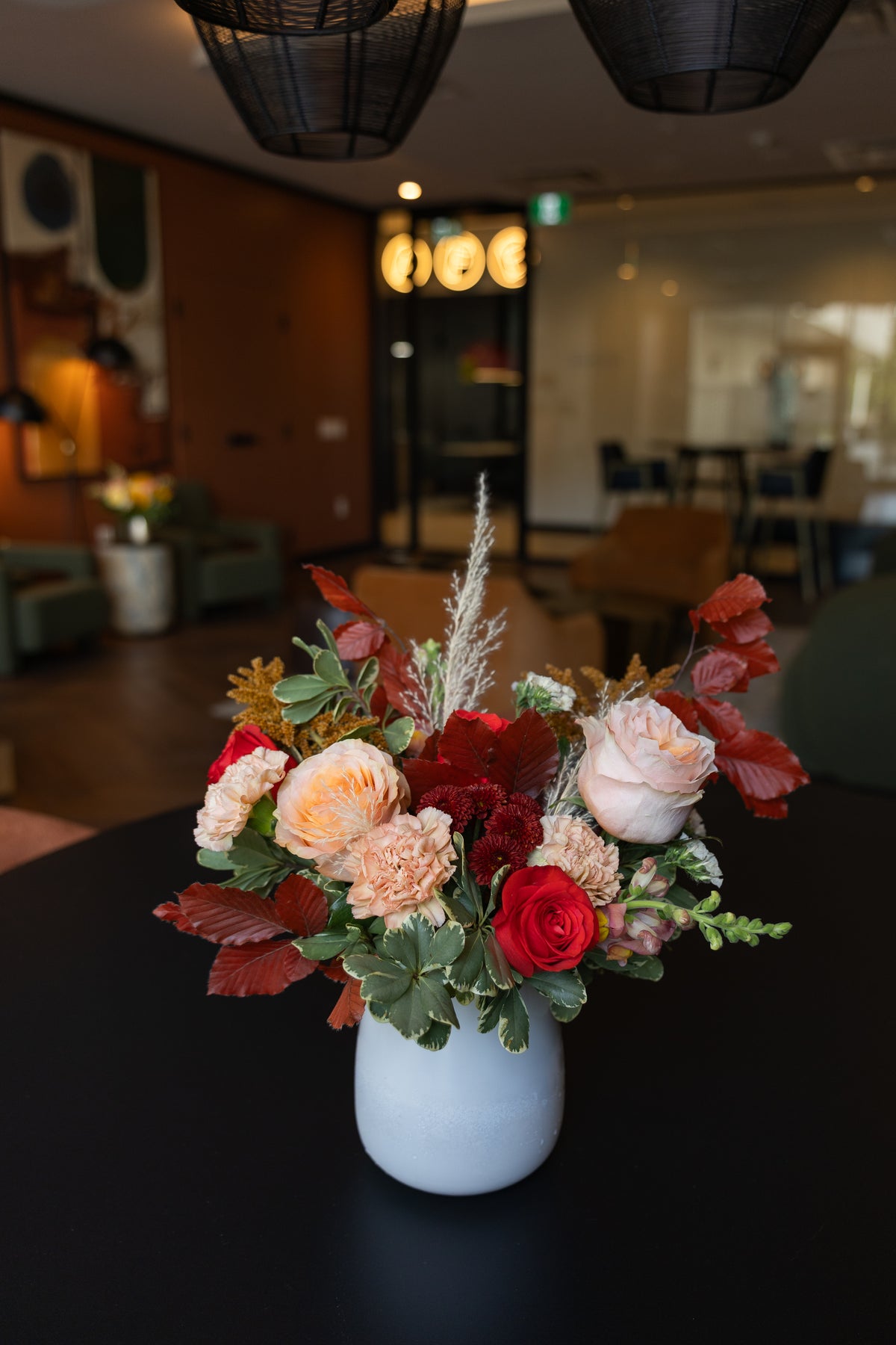 A vibrant floral arrangement featuring red and peach roses, green foliage, and dried grasses, presented in a matte white ceramic vase on a dark table with a blurred office lobby background.