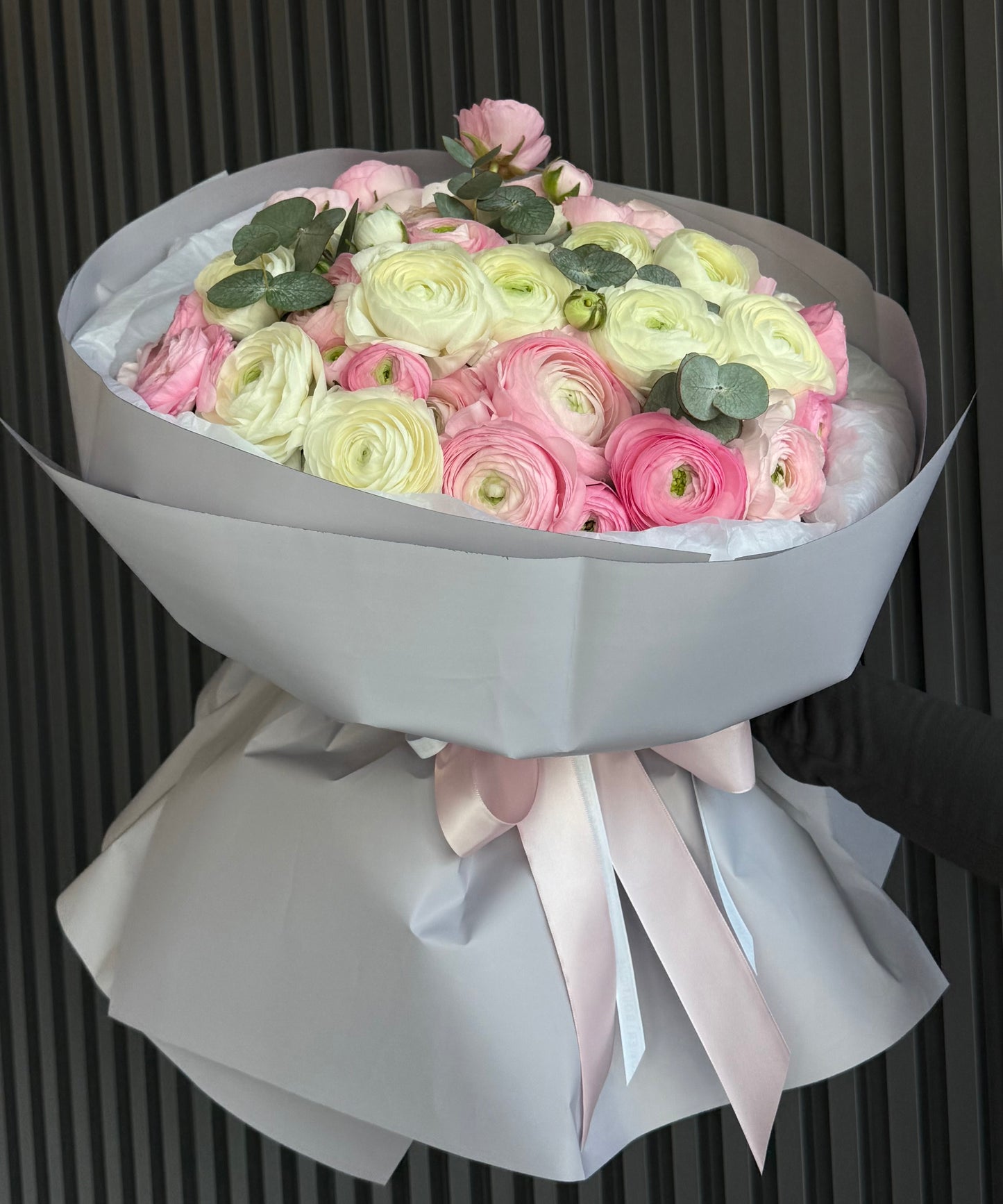 Bouquet of pink and white ranunculus wrapped in gray paper with a pink ribbon against a dark background.